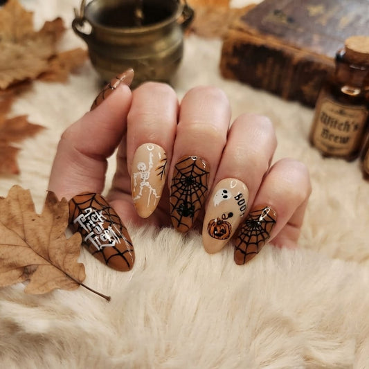 A hand with Halloween-themed manicure featuring brown spiderwebs, black spiderwebs, a skeleton, a pumpkin, and the words 'Spooky' and 'Halloween' on a beige faux-fur background with autumn leaves and witch potion bottles.