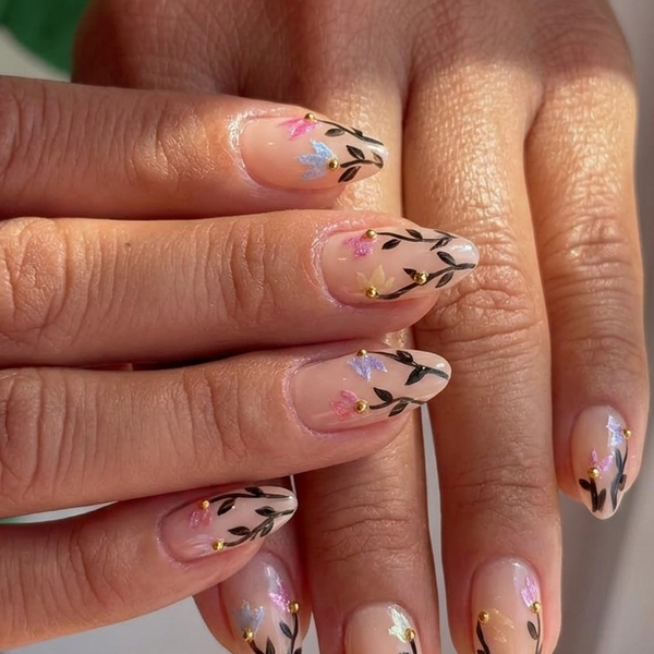 Close-up of a hand with manicured nails featuring a glossy nude-pink base, delicate black leafy vine patterns, and small pastel floral accents in pink, blue, and yellow.