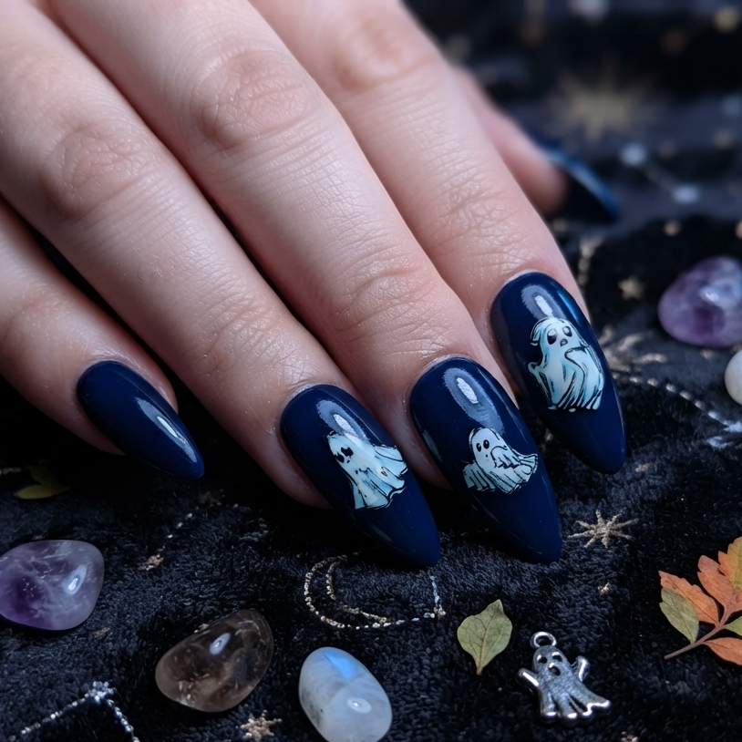A close-up of a hand with long almond-shaped nails painted in glossy dark navy blue, decorated with white ghost illustrations. The hand rests on a black fabric background printed with gold celestial motifs and surrounded by small decorative crystals and leaves.