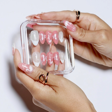 A pair of hands holding a clear plastic case containing pink and white press-on nails with a glossy finish, set against a clean white background.