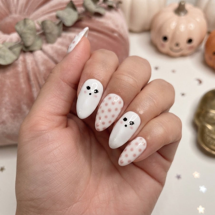 A hand with Halloween-themed nail art featuring white nails with black ghost faces and pale pink nails with pink polka dots, styled with a pink velvet pumpkin and small jack-o'-lanterns in the background.