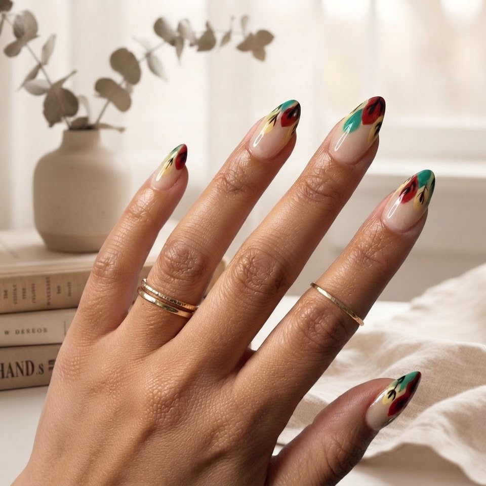 A close-up of a hand with almond-shaped nails painted in a glossy beige base with red, green, and yellow abstract accents, styled with gold rings in a bright, softly lit indoor setting.