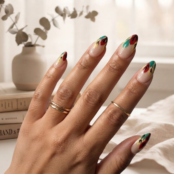 A close-up of a hand with almond-shaped nails painted in a glossy beige base with red, green, and yellow abstract accents, styled with gold rings in a bright, softly lit indoor setting.