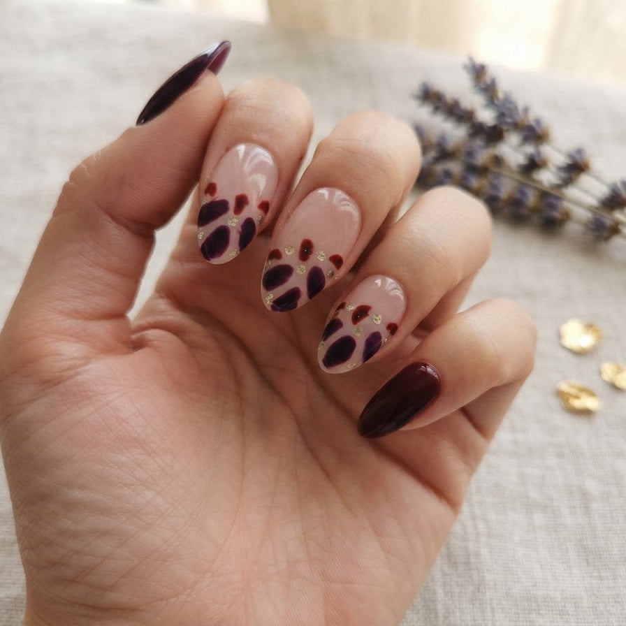 A hand with manicured nails featuring a glossy dark burgundy polish on the outer edges and a white base with black paw print designs on the inner nails, resting on a textured beige fabric with dried lavender and gold flakes in the background.