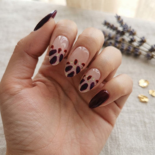 A hand with manicured nails featuring a glossy dark burgundy polish on the outer edges and a white base with black paw print designs on the inner nails, resting on a textured beige fabric with dried lavender and gold flakes in the background.