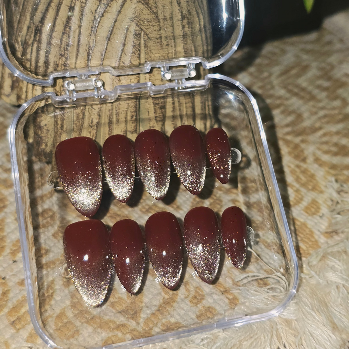 A set of ten false press-on nails with a metallic silver glitter cat-eye finish, displayed in a clear plastic case on a textured beige surface.