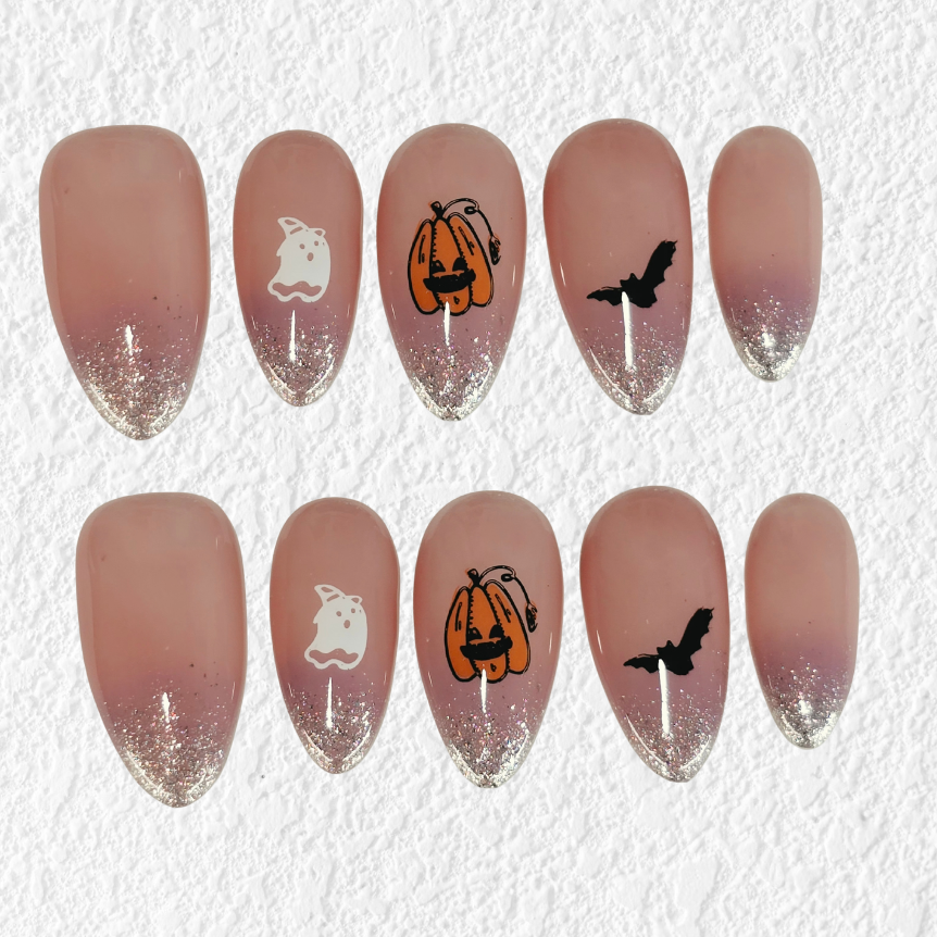 A set of ten press-on nails with a dusty rose to silver glitter gradient, decorated with Halloween motifs including pumpkins, ghosts, and bats on a textured white background.