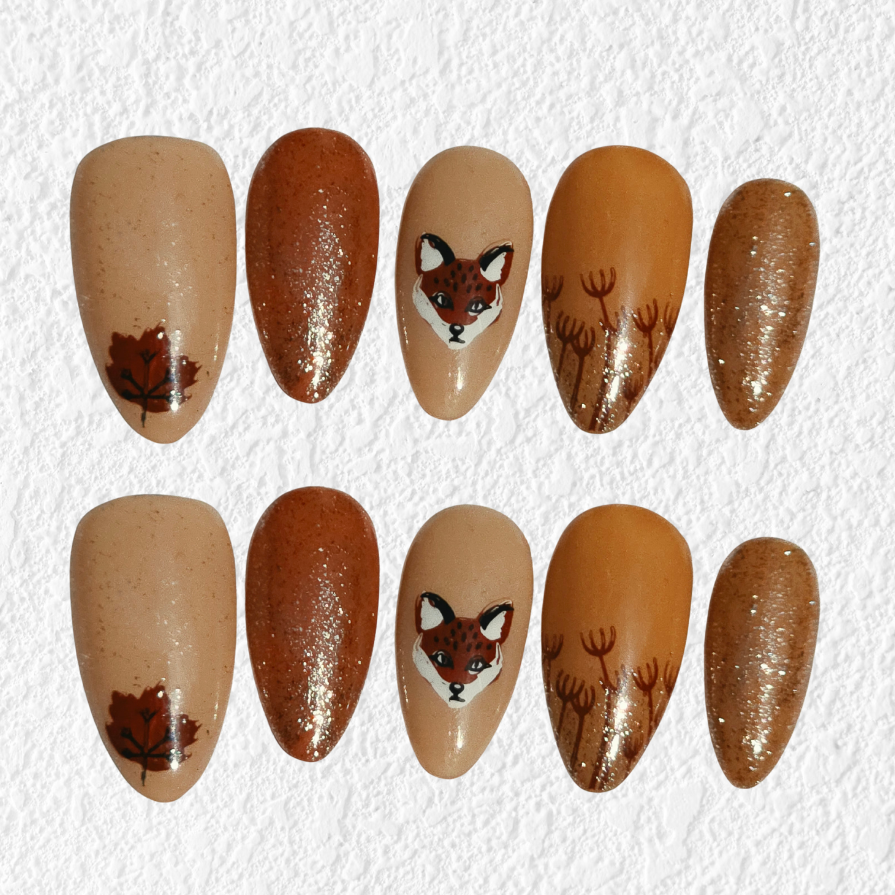 Ten almond-shaped press-on nails arranged in two rows on a textured white background. The nails feature autumn and woodland designs, including a maple leaf, a fox head, and wildflower motifs in shades of brown, tan, and glitter.