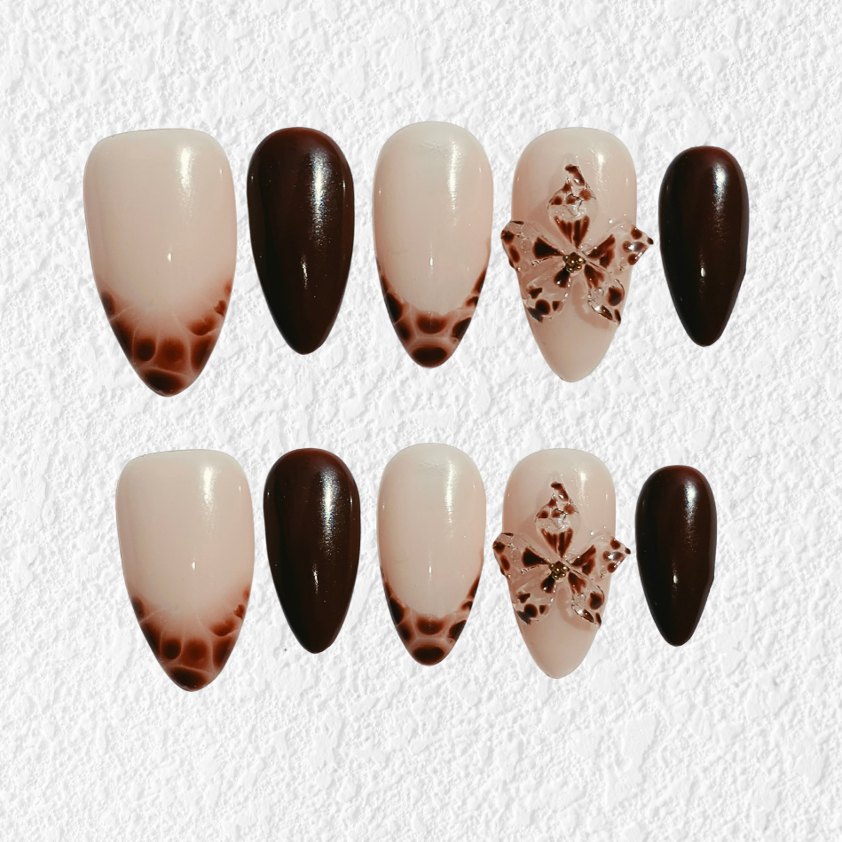 A set of 10 press-on nails arranged in two rows on a textured white background. The design features a mix of solid dark brown, glossy dark brown, cream-to-brown gradient tips, and one nail with a brown snowflake pattern on a cream base.