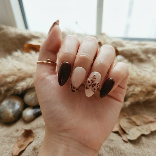 A hand with almond-shaped nails styled in a dark brown and beige bohemian manicure, resting on a textured beige blanket with stones and dried leaves.