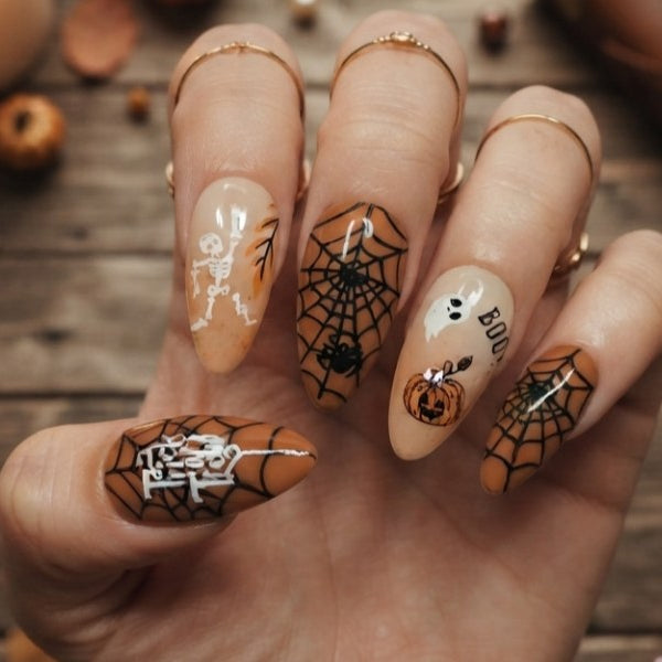 A hand with Halloween-themed nail art, featuring spiderwebs, pumpkins, and skeletons on a rustic wooden background with autumn leaves.